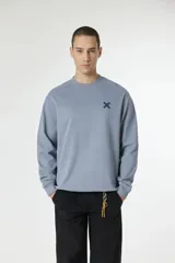 Buzo de cuello redondo en color azul grisáceo, con diseño de manga larga y puños acanalados. Presenta un pequeño logo bordado en forma de cruz en el pecho.