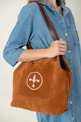Bolso tipo tote de gamuza color chocolate, con bolsillo frontal externo que presenta un diseño bordado circular con una flor de lis estilizada. Posee correas de hombro de cuero marrón oscuro.