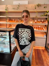 Remera oversize de algodón negro, con diseño gráfico frontal que incluye la leyenda "COFFEE CLUB" y una ilustración estilo retro de una mujer bebiendo café.