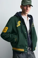 Cazadora tipo varsity de corte relajado confeccionada en tejido efecto ante color verde oscuro. Presenta cuello solapa, cierre frontal con botones a presión y puños y bajo acanalados con rayas en contraste. Incluye parches bordados en color amarillo en el frente y mangas.
