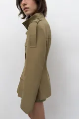 Blazer entallada color beige con cuello subido, manga larga con hombreras y trabillas en los hombros. Presenta bolsillos de plastrón en el delantero, detalle de solapas en el pecho y cierre frontal con botones a presión.