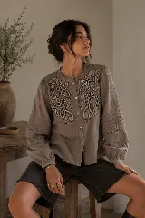 Camisa de color marrón topo con bordados ornamentales en el frente, cuello redondo, cierre frontal de botones y mangas largas con detalles de alforzas, calados y puños elásticos.