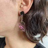 Par de aros colgantes con base negra, cadena plateada y dije transparente con dibujo de ojo rosa.