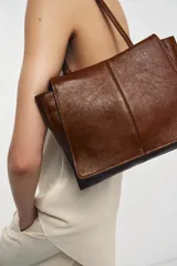 Bolso tipo mini shopper confeccionado en piel con acabado brillante. Presenta un diseño de doble compartimento con cierre de botón imantado y asa de hombro ajustable.