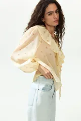 Blusa de gasa arrugada de corte holgado, en tono amarillo claro con estampado floral. Cuenta con escote en V profundo, un amplio volante ribeteado con encaje alrededor del cuello y cordones finos para atar al frente. Mangas largas con puños elásticos.