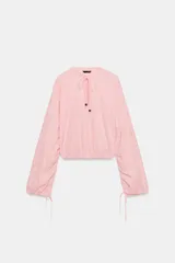 Blusa de manga larga con estampado de cuadros vichy en tonos rosado y blanco. Presenta cuello redondo con volante, cordón ajustable en el escote con apliques decorativos, y puños fruncidos con tiras para atar.