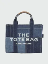 Cartera tipo tote bag Marc Jacobs modelo The Small Traveler Tote, confeccionada en denim con diseño de paneles en distintos tonos de azul. Cuenta con asas superiores, correa de hombro ajustable y extraíble, y el texto "THE TOTE BAG MARC JACOBS" estampado en la parte frontal.