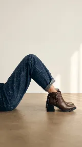 Bota corta de cuero marrón con diseño de paneles laterales texturizados. Presenta un taco ancho cuadrado en color negro y suela de plataforma baja, ofreciendo un estilo moderno y estable.