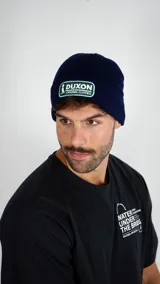 Gorro tipo beanie de punto color azul marino, con parche rectangular bordado en el frente que incluye el logo y nombre de la marca Duxon.