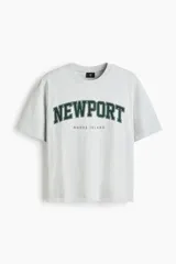 Remera de algodón gris jaspeado con cuello redondo y mangas cortas. Presenta un estampado frontal con la inscripción 'NEWPORT' en tipografía universitaria verde oscuro y 'RHODE ISLAND' debajo.