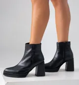 Bota corta de cuero negro con diseño combinado, presentando una textura lisa en la parte frontal y una textura tipo reptil en el talón. Cuenta con taco cuadrado alto, plataforma delantera y cierre lateral interno.