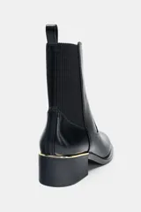 Botín de cuero sintético negro con caña elástica lateral, tirador trasero y detalle de aplique metálico dorado en la base del tacón cuadrado.
