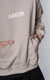 Hoodie oversize color gris topo, confeccionado en rústico de algodón, con capucha forrada, bolsillo canguro y costuras expuestas en mangas y ruedo. Presenta estampa gráfica en la espalda con la palabra AMOR y diversos motivos románticos.