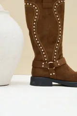 Bota alta de gamuza color marrón, con suela plana y gruesa, puntera redondeada y caña alta decorada con costuras en relieve y tachas plateadas. Cuenta con una tira con hebilla y argolla metálica en la base del empeine.