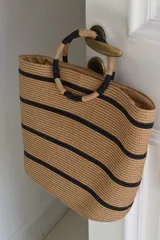 Bolso tipo tote confeccionado en material tejido color tostado con diseño de rayas horizontales negras y asas de mano circulares rígidas envueltas.
