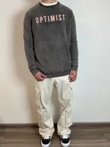 Buzo de cuello redondo con efecto lavado, color gris oscuro. Presenta estampado frontal con la palabra 'OPTIMIST' en letras claras y 'DUXOR COLLECTION' debajo en letras oscuras. Posee puños y ruedo acanalados.