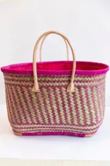 Bolso tipo tote de raffia natural con diseño tejido, interior forrado en tela fucsia y doble asa de cuero color beige. Tiene base rectangular y detalles de cuero en las esquinas inferiores.