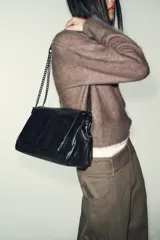 Bolso de hombro con solapa, confeccionado en material sintético con acabado brillante y efecto envejecido. Presenta un diseño estructurado con costuras decorativas, tres compartimentos internos, uno de ellos con cierre de cremallera, y asa de cadena metálica. Se cierra mediante un imán oculto.