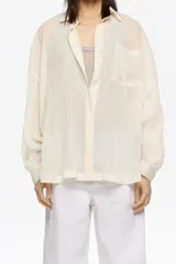 Camisa larga de corte oversize color crema, confeccionada en tejido plisado. Presenta cuello camisero, botones frontales y bolsillo plastrón en el pecho.