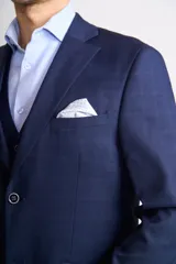Traje de tres piezas color azul marino con textura sutil. Incluye saco entallado con solapas clásicas, chaleco abotonado y pantalón de corte recto. Se complementa con una camisa celeste de cuello italiano y pañuelo de bolsillo blanco.
