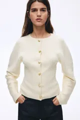 Saco ajustado en punto acanalado color beige, con cuello redondo, cierre frontal con botones jaspeados, hombros caídos y mangas largas con puños ceñidos.