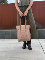 Bolso tipo tote bag de símil cuero color rosa pálido, con asas dobles y detalles de costura en color beige claro. Presenta un bolsillo frontal rectangular aplicado y herrajes metálicos.