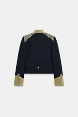 Chaqueta de estilo militar en color negro, con cuello subido y puños en terciopelo color verde oliva. Presenta detalles de alamares dorados en el frente y hombros, bolsillos con solapa y cierre frontal con botones metálicos en relieve.