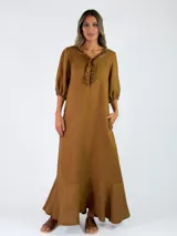 Vestido largo de lino color marrón tostado, con mangas 3/4 abullonadas, escote en V con volados y botones frontales. Cuenta con bolsillos laterales y detalle de volado en el dobladillo inferior.