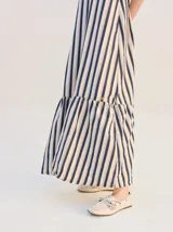 Vestido largo de algodón denim con diseño de rayas verticales en color azul marino y beige. Cuenta con tirantes finos ajustables con lazos en los hombros, escote recto y corte suelto con un volante en el bajo.