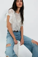Top corto blanco confeccionado en encaje de nailon con diseño floral transparente, escote redondo y mangas cortas holgadas con terminación festoneada.