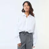 Camisa de popelina blanca con cuello clásico, cierre frontal de botones y mangas largas abullonadas con puños ajustados.
