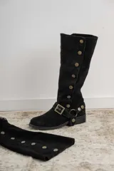 Botas altas de gamuza negra con caña alta, detalle de tachas metálicas laterales y correa con hebilla en el tobillo.