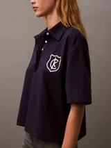 Chomba azul marino de punto de algodón con cuello clásico, tapeta con botones ocultos y mangas cortas. Presenta un corte holgado y un escudo bordado con el logo de Calvin Klein en el pecho.