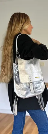 Mochila de cuero color plateado con diseño de solapa frontal, cierre de hebilla decorativa y correas ajustables. Cuenta con herrajes metálicos y una placa con logo en la parte frontal.