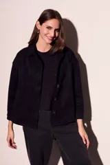 Sobrecamisa negra de estilo shacket, confeccionada en tejido de gamuza sintética. Presenta cuello camisero, cierre frontal de botones y dos bolsillos de parche con solapa en el pecho. El calce es oversize y las mangas son largas.
