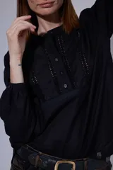 Camisa blanca de algodón con pechera decorada con alforzas y puntillas. Presenta cierre frontal con botones, mangas tres cuartos con frunces y detalles de puntilla, y un diseño de corte holgado con ruedo redondeado.