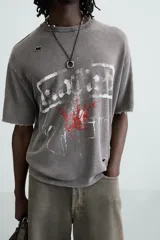 Camiseta gris de punto con efecto lavado y roturas, corte "boxy fit", cuello redondo y manga corta. Presenta un estampado frontal en blanco y rojo con diseño abstracto.