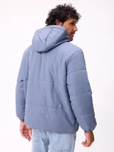 Campera puffer Rusty de color azul, con capucha desmontable, cierre frontal con botones a presión y bolsillos laterales. Presenta un parche con el logo de la marca en el pecho.