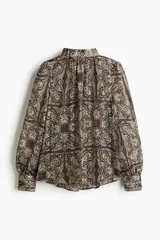 Blusa de tejido transparente con cuello elevado y botones forrados en la nuca. Presenta mangas largas con puños abotonados y un bajo levemente redondeado con estampado paisley.