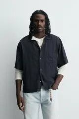 Camisa de algodón negro, de corte relajado (relax fit), con cuello bowling, manga corta y bolsillo plastrón en el pecho. Cierre frontal con botones a presión.