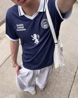 Camiseta de fútbol de Escocia de manga corta, color azul marino con diseño a cuadros sutiles, cuello en V y cuello camisero blanco con líneas azules. Presenta detalles estampados en el frente, incluyendo el escudo de la Asociación de Fútbol de Escocia, un león rampante y el texto 'Scottish Football Association'.