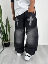 Pantalón de corte baggy en color negro con efecto lavado. Presenta parches decorativos en los bolsillos traseros con diseño de cruces y calaveras con bordes deshilachados.