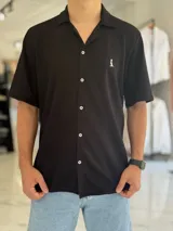 Camisa de manga corta color negro, confeccionada en tejido texturizado con cuello abierto tipo cubano, botones blancos frontales y pequeño bordado blanco en el pecho.