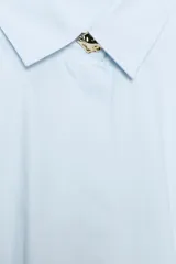 Camisa de popelín color celeste, con cuello solapa, manga larga y cintura entallada con pliegues. Presenta cierre frontal con botones ocultos por solapa.