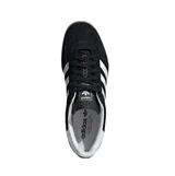 Championes Adidas Gazelle Indoor de diseño clásico, confeccionados en gamuza negra con las icónicas tres tiras laterales en blanco. Presentan el nombre del modelo en letras doradas, talón en contraste blanco y suela de caucho color caramelo.