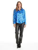 Camisa de satén de corte holgado con cuello camisero y cierre frontal de botones metálicos. Presenta un estampado de cuadrados negros sobre fondo azul vibrante, mangas largas con puños abotonados y un acabado brillante característico del satén.