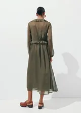 Vestido midi color verde militar, confeccionado en tejido fluido de poliéster. Presenta un diseño evasé, escote de pico con volantes, y mangas largas con puños elásticos. Incluye detalles de volantes en el cuello y forro interior.