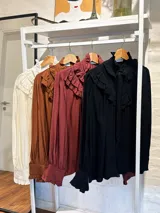 Blusa de color marrón con diseño romántico, cuello alto con lazo ajustable y volados tipo shabot en el frente. Presenta mangas largas abullonadas con puños ajustados y una textura suave y liviana.