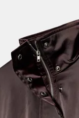 Cazadora satinada de color negro, con cuello subido, manga larga y bolsillos frontales de plastrón con cordones decorativos. Cuenta con bajo elástico y cierre frontal con cremallera y botones a presión oculta.