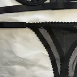 Conjunto de lencería negro de tiras elásticas con arandelas metálicas, compuesto por un soutien tipo arnés y una bombacha de encaje y malla transparente.